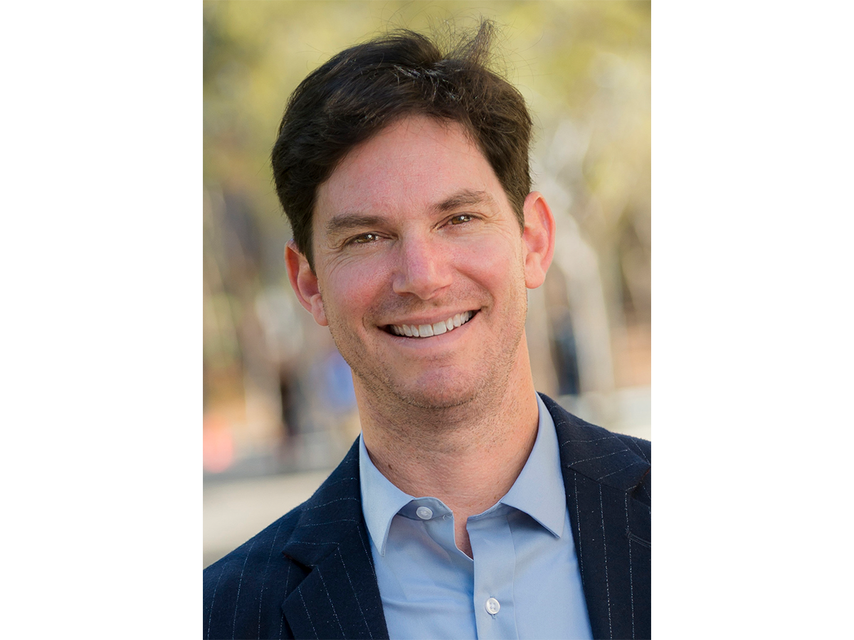 Professor Seth Cohen smiling for camera in pale open neck shirt and jacket