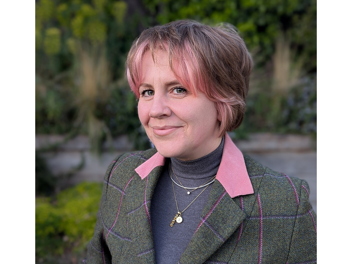 Headshot of Zoe smiling with pink hair and suit jacket and polarneck