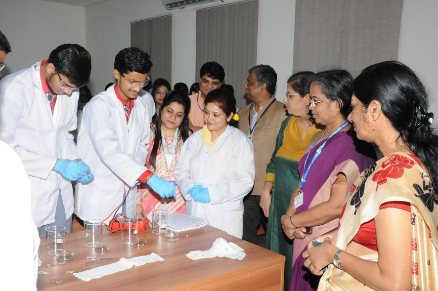 Two young men wearing safety glasses and white coats conduct an experiment using coins