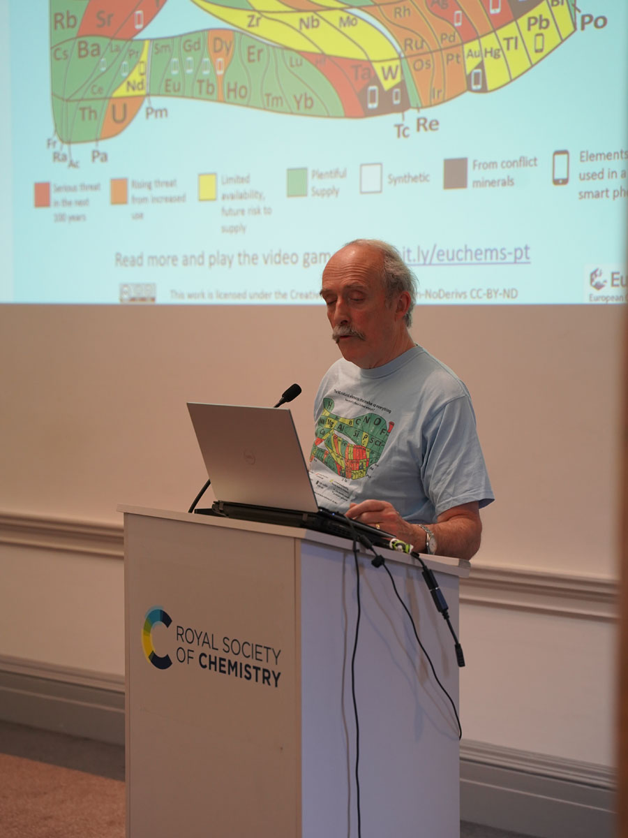 David Cole-Hamilton, wearing a T-shirt featuring the scarcity of the elements periodic table, speaks from behind a lectern. The scarcity of the elements periodic table is visible on a screen behind him.