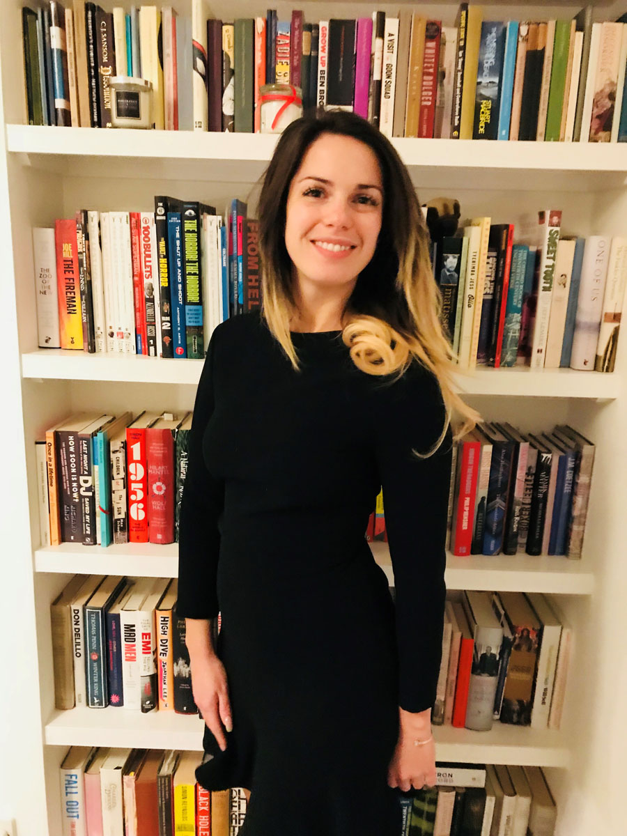 Kristen stands in her home in front of a ceiling-high bookshelf. She is smiling at the camera and wearing a black dress. Kristen stands in her home in front of a ceiling-high bookshelf. She is smiling at the camera and wearing a black dress.