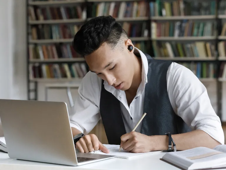 a man writing in a book in front of a laptop