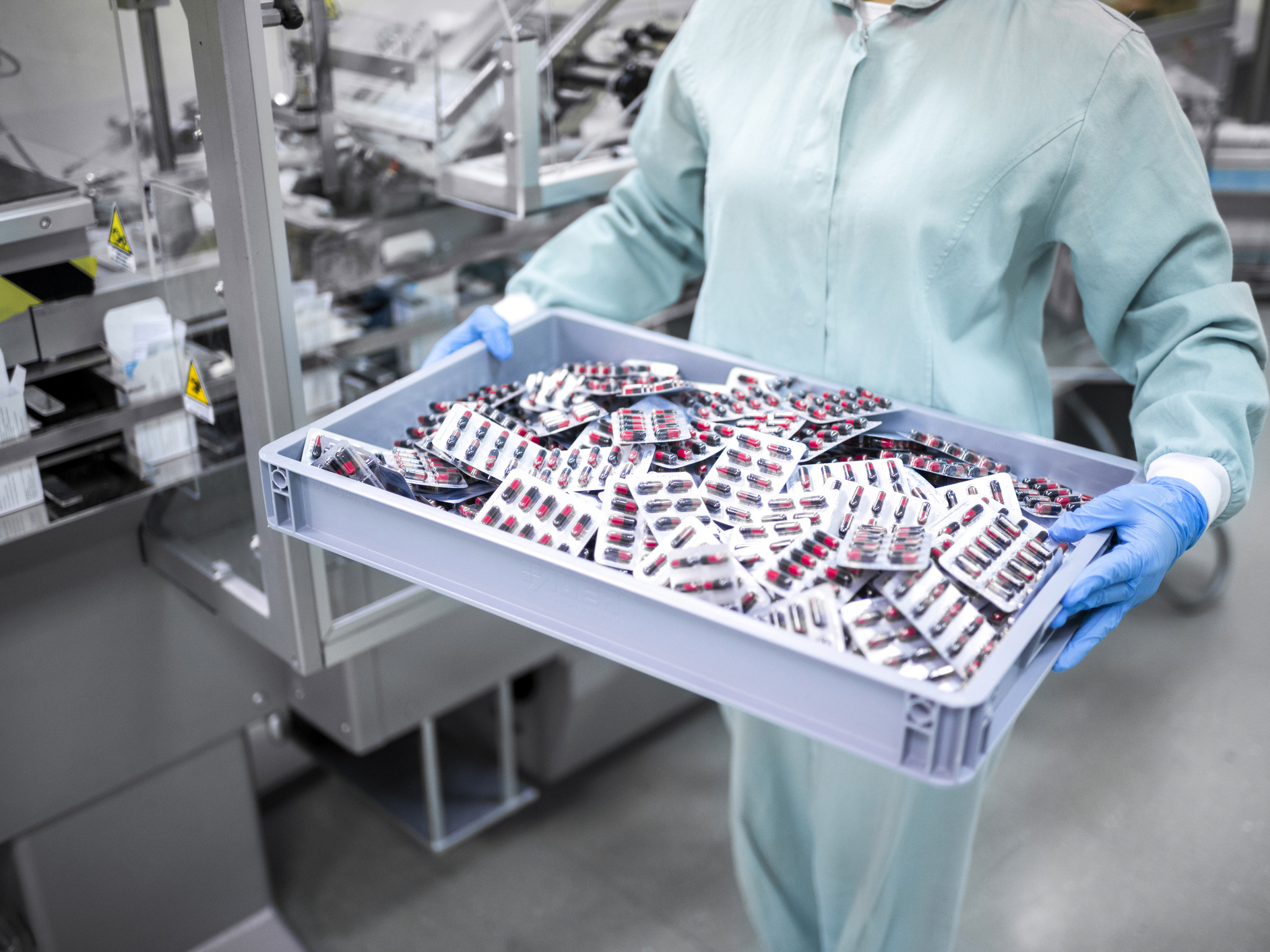 A worker in protective gear carefully carries a container filled with blister-packaged capsules