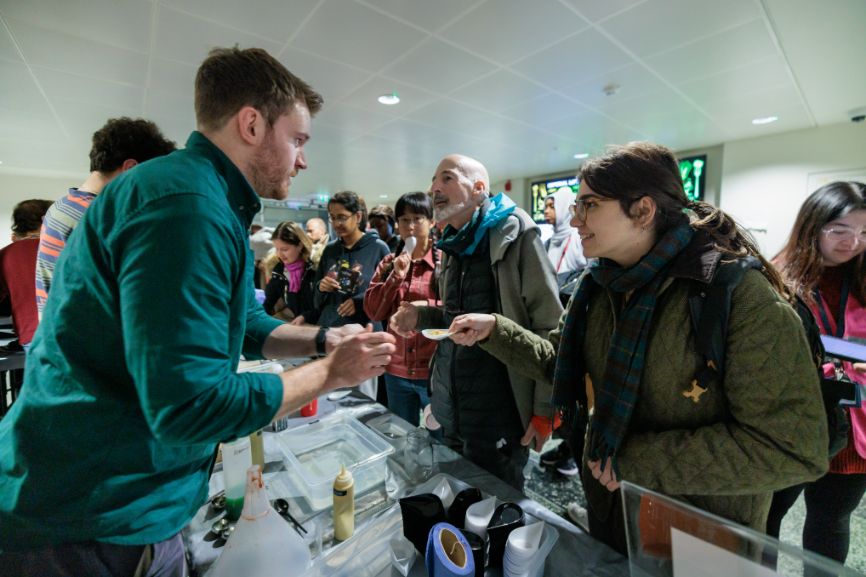 A man at a stall talks to a woman holding a spoon with a dessert on it.