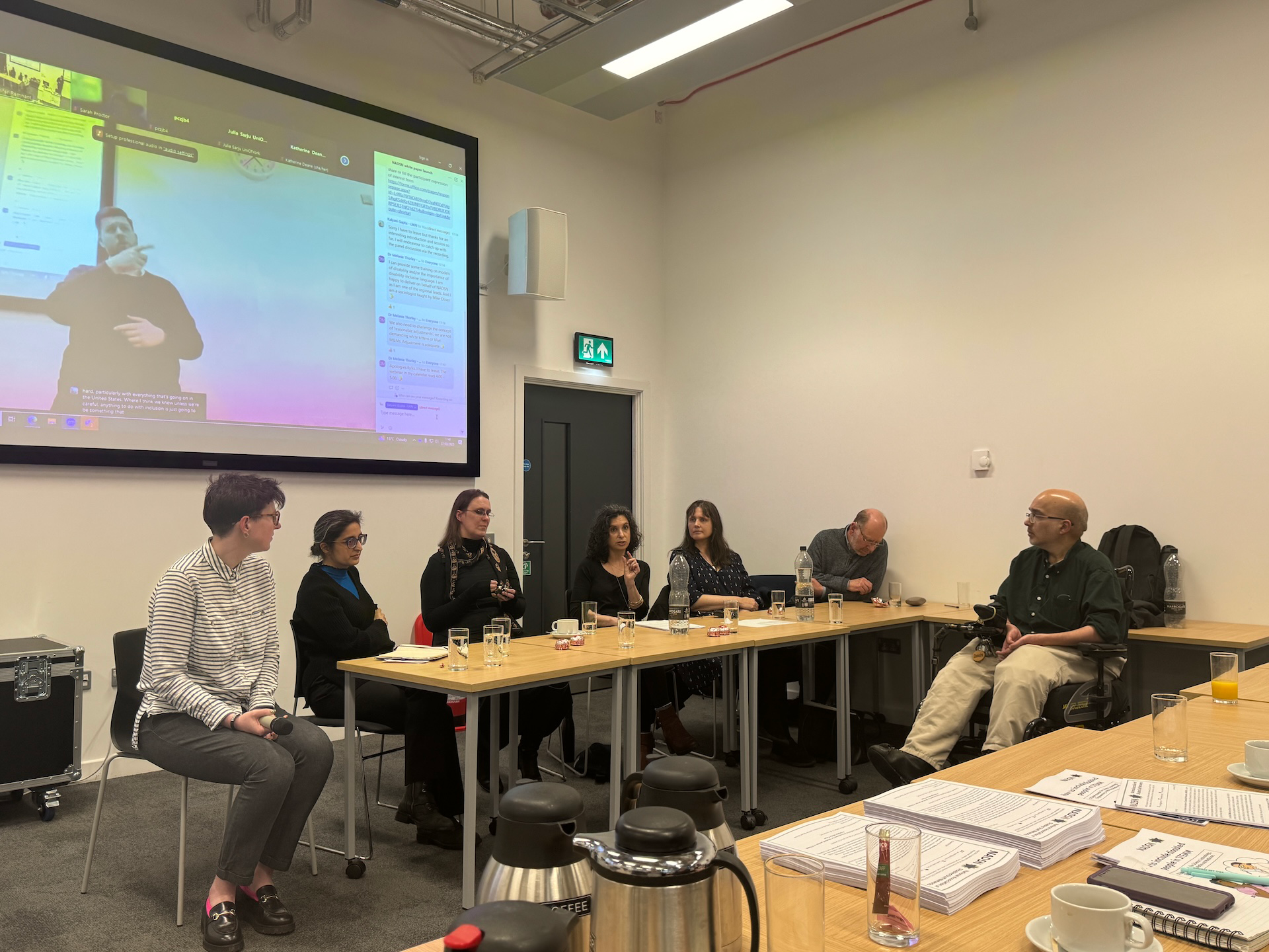 Professor Jennifer Leigh sitting around a table with six others in discussion, a person is performing sign language on a screen on the wall