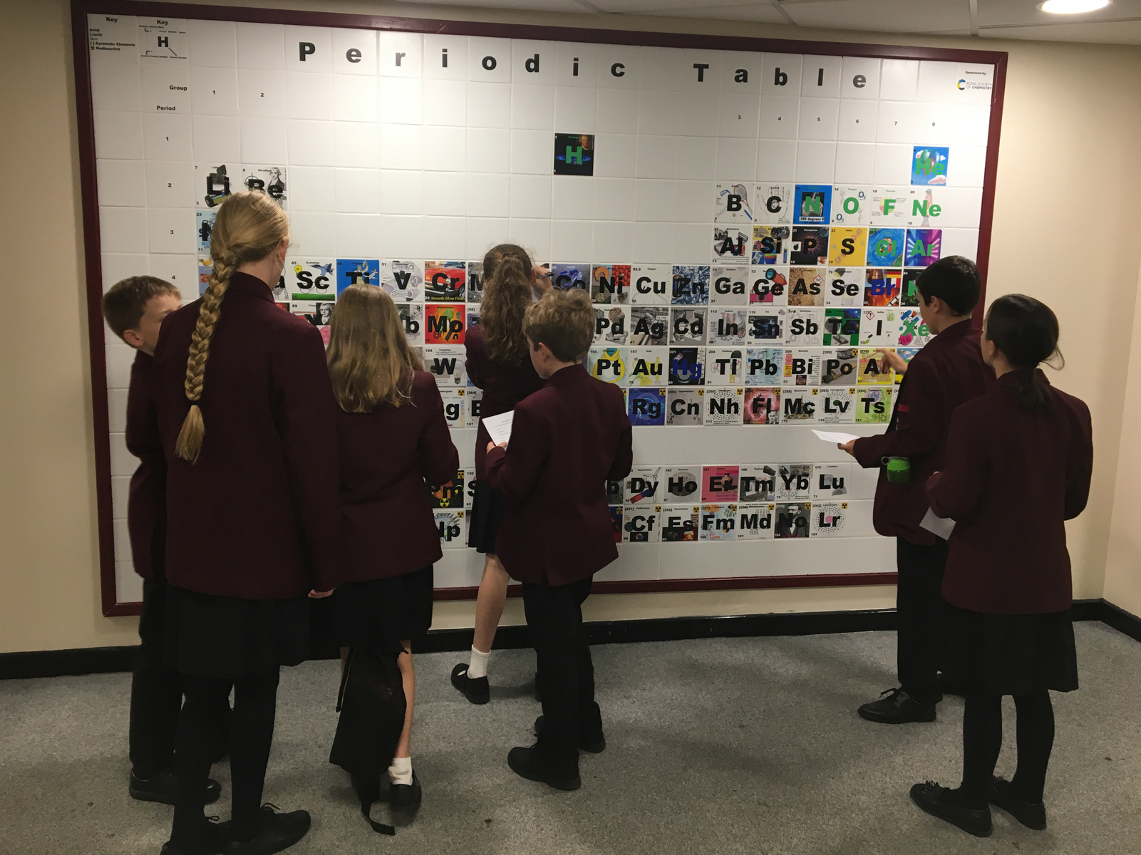 Schoolchildren with their backs to the camera looking at the large ceramic periodic table on the wall and taking notes.