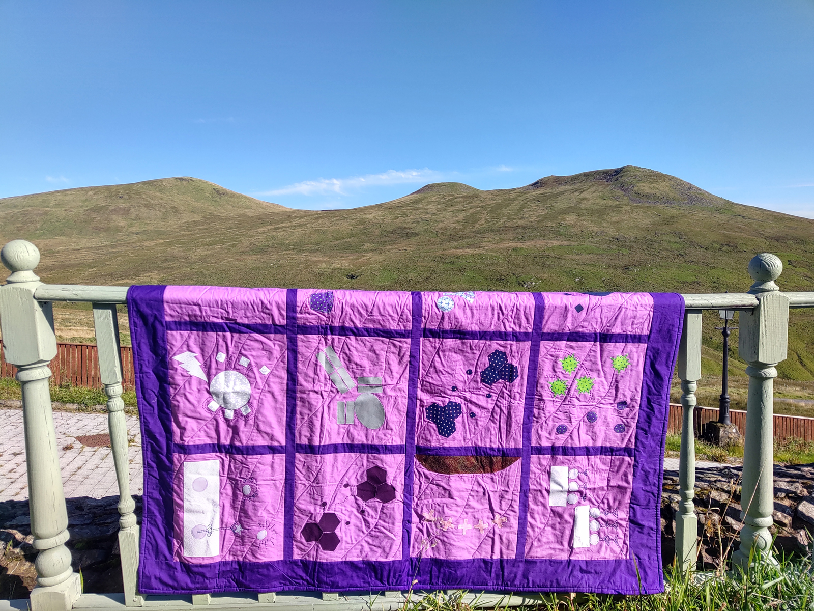 Purple quilt displayed on a railing with mountains in the background. The quilt is embroidered with chemical and biological symbols.