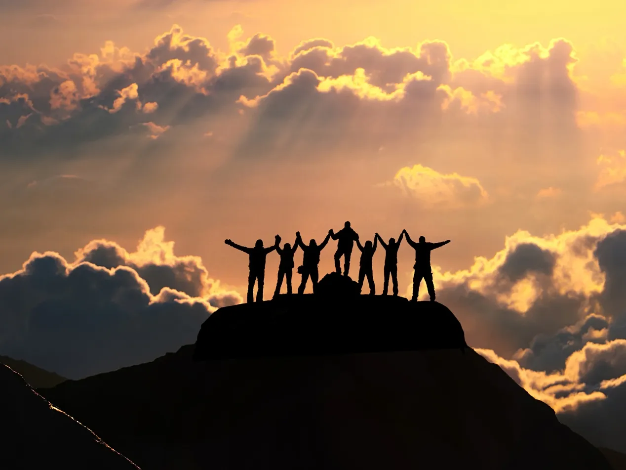 Silhouette of a group standing on top of mountain with arms raised with the sun setting behind them