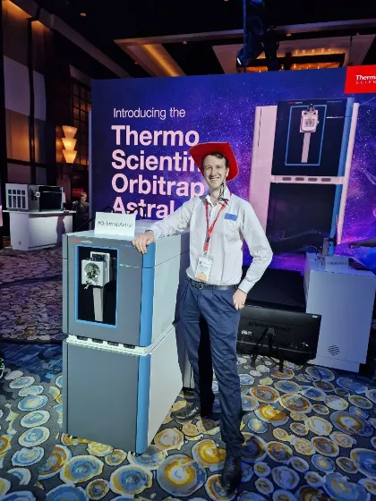 One of the male members of the Orbitrap Astral Team standing next to a large machine at an event