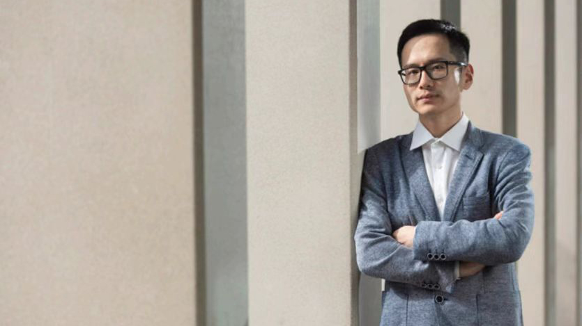 Dr Guanjie He in a suit and glasses with his arms crossed, looking at camera