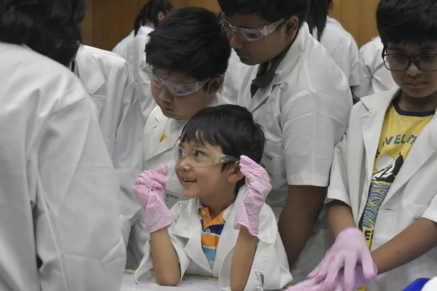 An excited young boy wearing safety goggles and gloves watches an experiment with other children