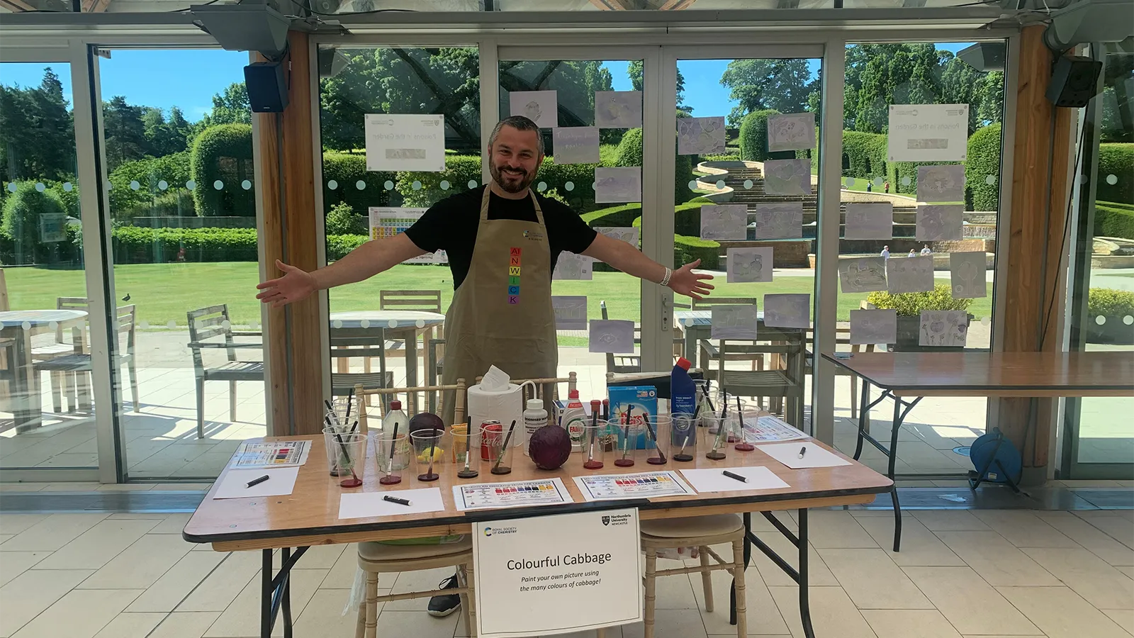 Dr Graeme Turnbull standing in an apron with open arms, showing his experiments laid out on table in front of him