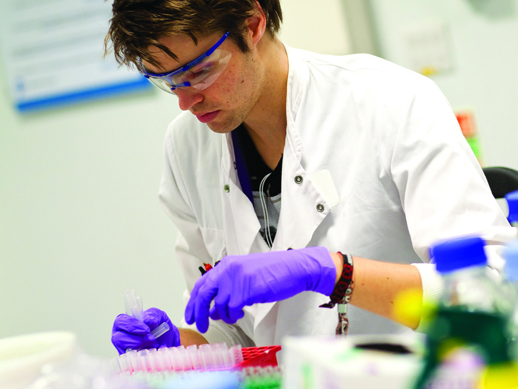 A man in a lab coat and gloves examines a specimen in a laboratory