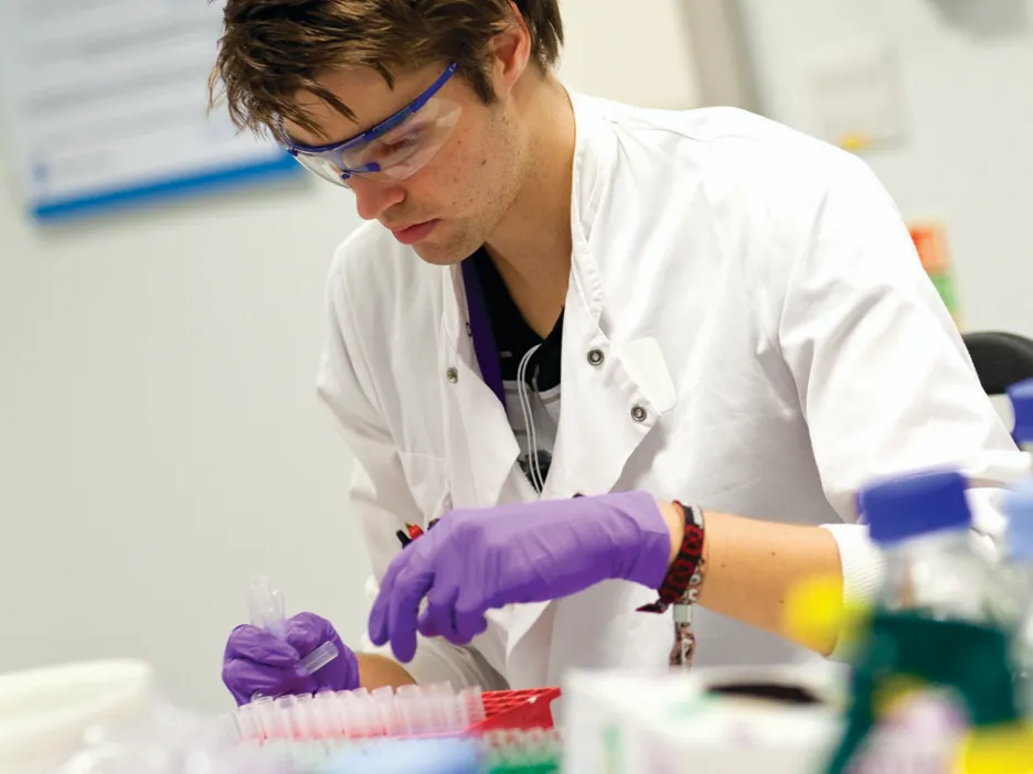 A man in a lab coat and gloves examines a specimen in a laboratory