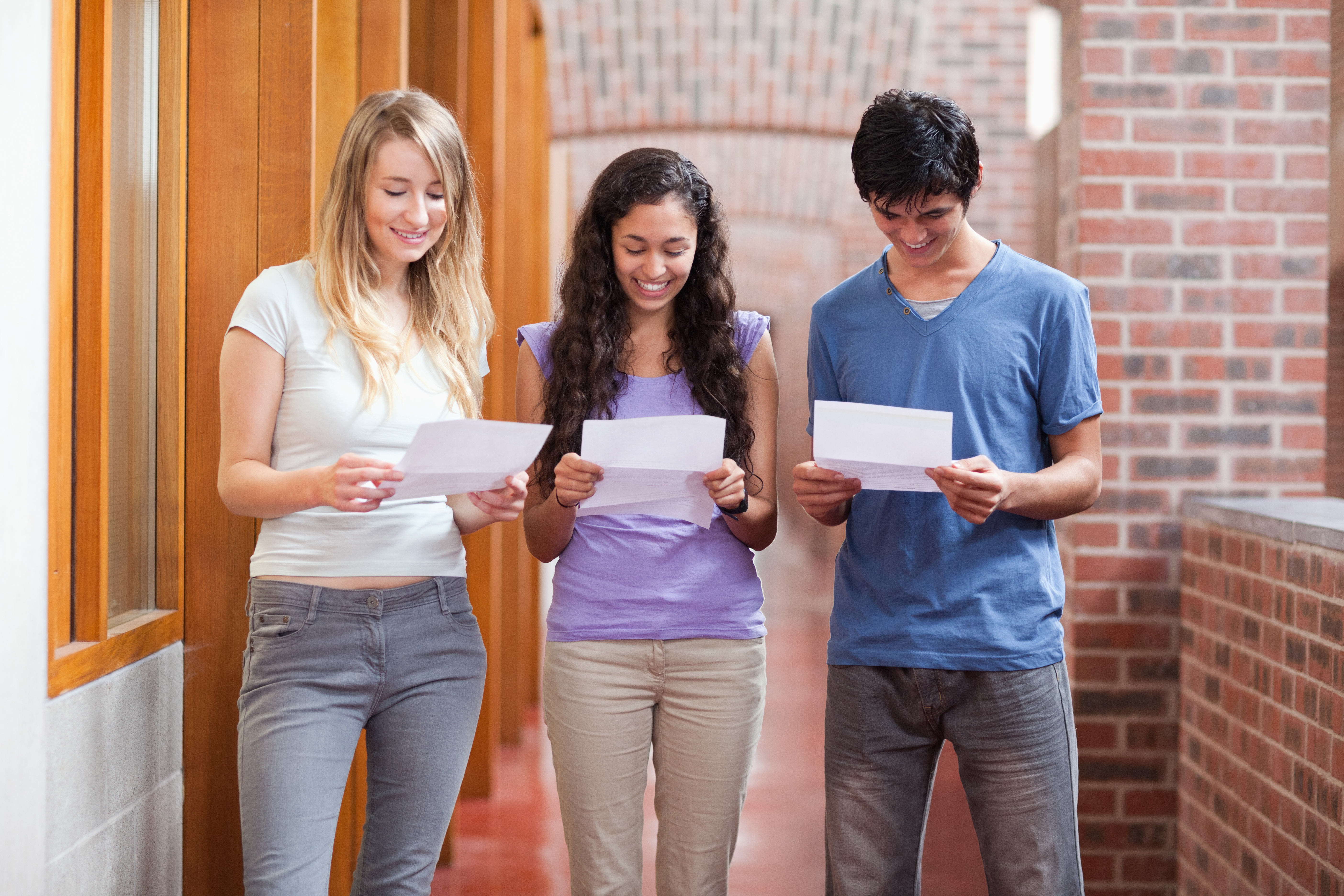 Three high school students, two female and one male, stand together looking down at pieces of paper with their exam results on them Three high school students, two female and one male, stand together looking down at pieces of paper with their exam results on them