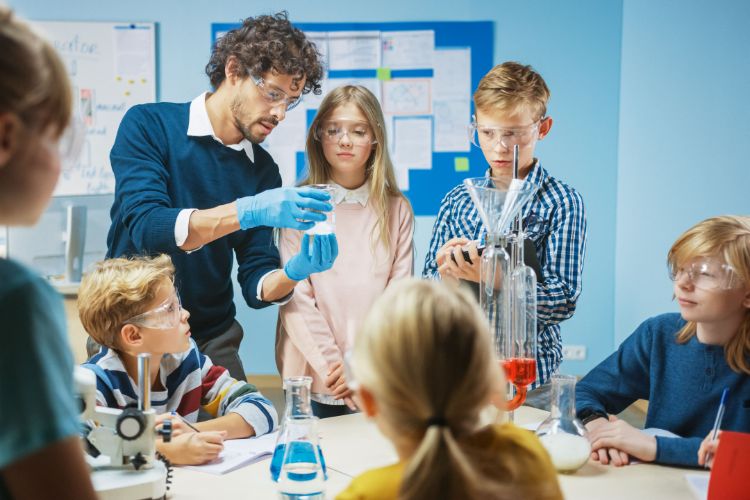 A science teacher teaches five schoolchildren in a classroom, while wearing safety goggles and doing something with glassware A science teacher teaches five schoolchildren in a classroom, while wearing safety goggles and doing something with glassware