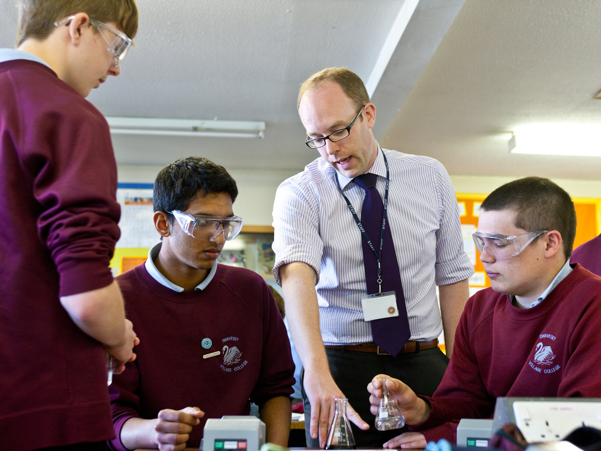 Three male school pupils in their early teens, wearing safety goggles and burgundy jumpers sit either side of a tall science teacher, who is wearing glasses, a shirt and tie and a lanyard Three male school pupils in their early teens, wearing safety goggles and burgundy jumpers sit either side of a tall science teacher, who is wearing glasses, a shirt and tie and a lanyard