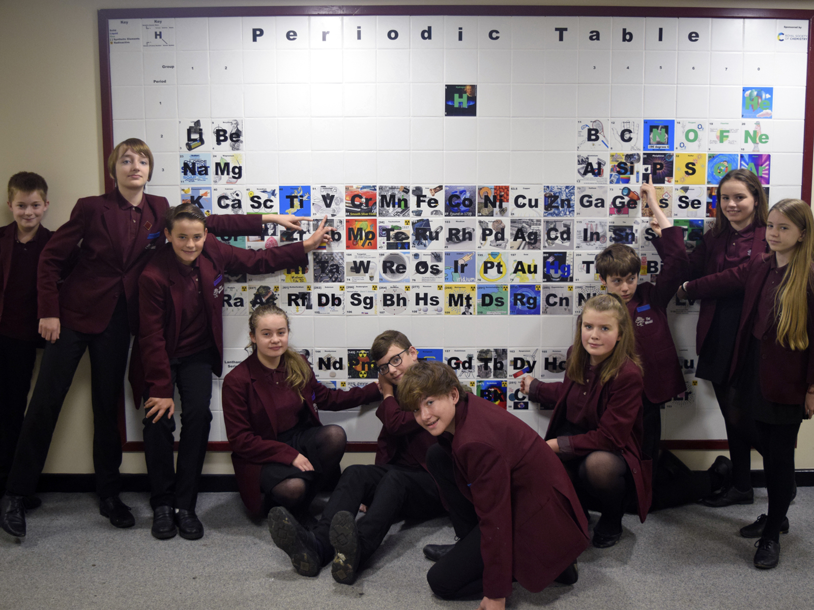 School children in burgundy blazers, some sitting some standing, smile at the camera and point at a large and colourful ceramic periodic table on the wall