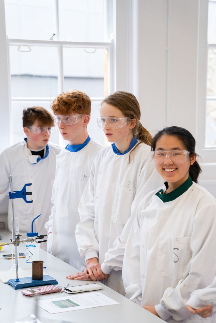 Four students, donning white coats and safety goggles, work away in the lab