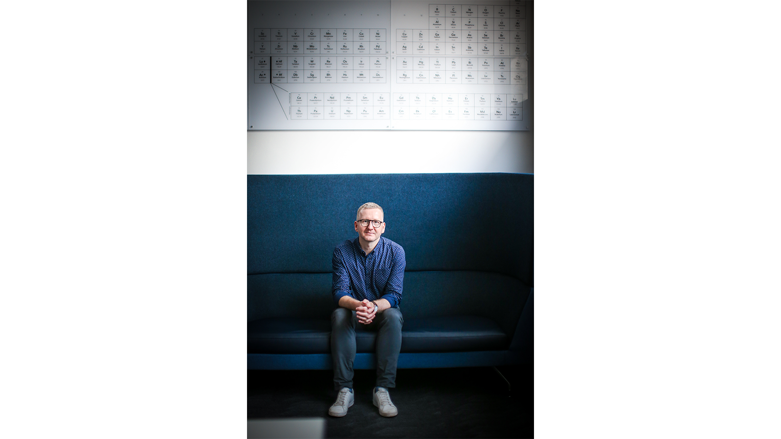 One of the team sitting on blue sofa with periodic table on the wall above him