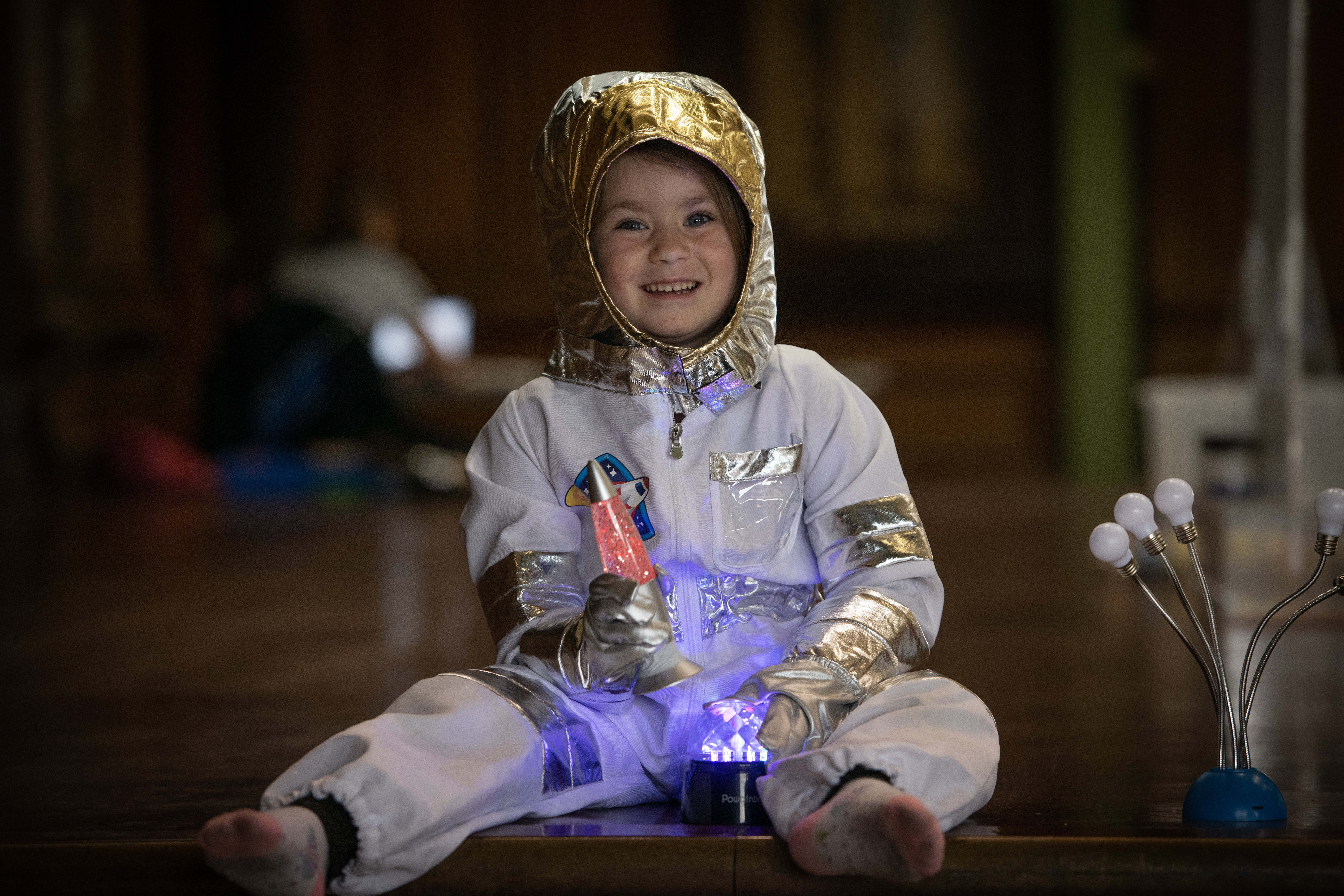 A little girl in an astronaut costume smiles as she holds a lava lamp and sits in front of a little light