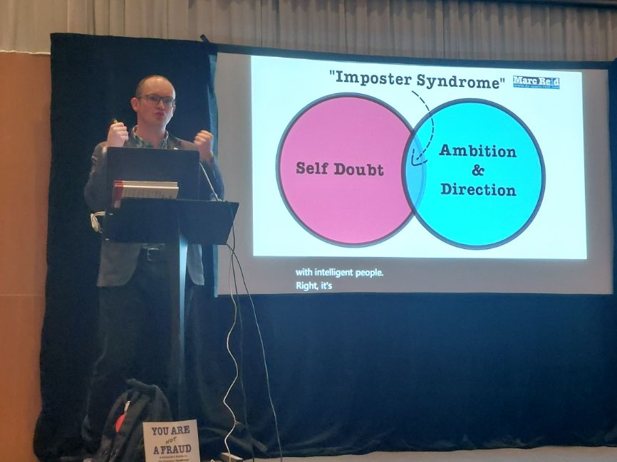 Dr Marc Reid gives a presentation to the group, who are out of shot. The screen behind him says "Imposter syndrome" and underneath is a Venn diagram with "Self doubt" on one side a