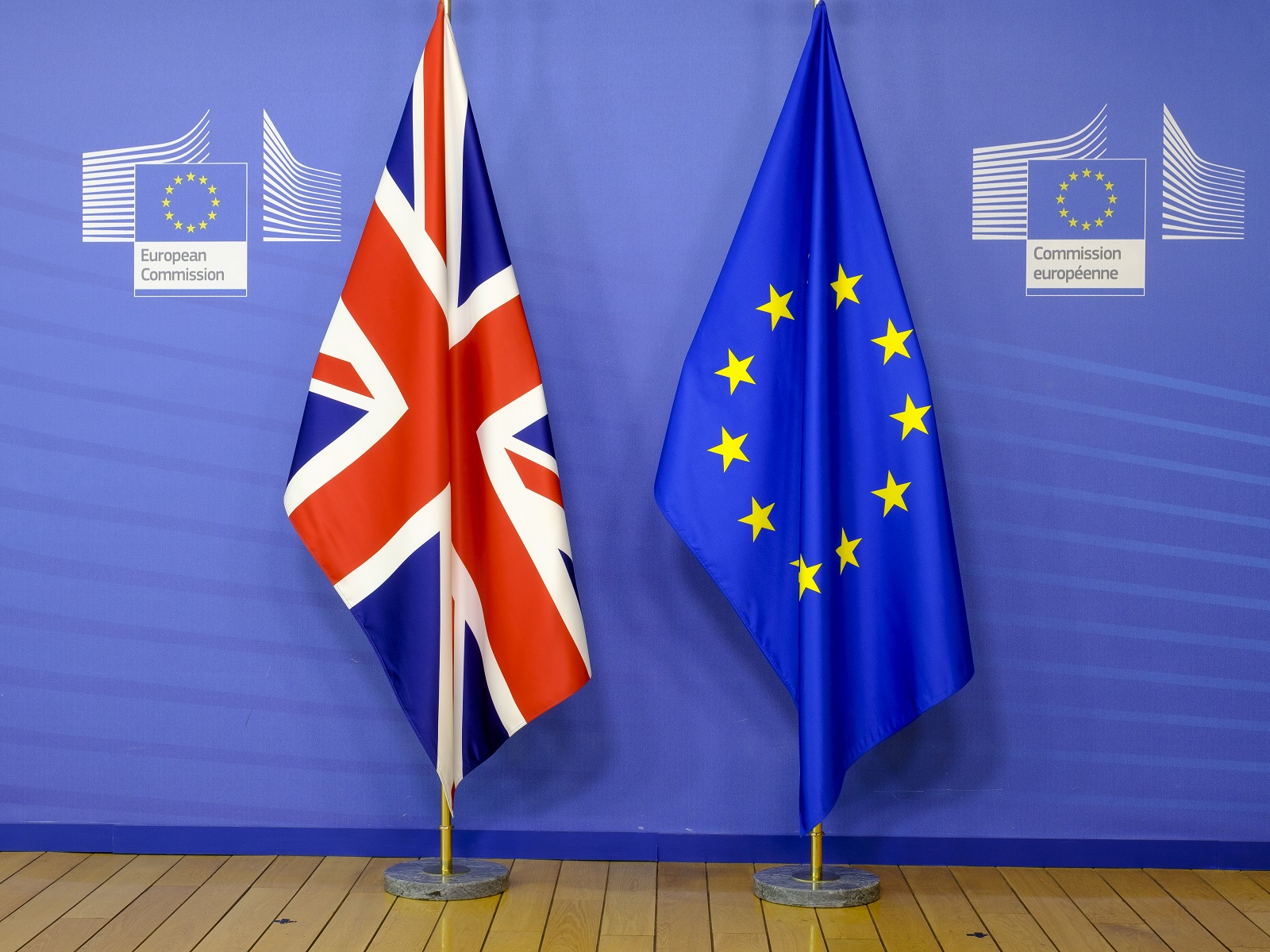 A Union Jack and an EU flag droop from flagpoles in front of a blue European Commission background and above a wooden floor