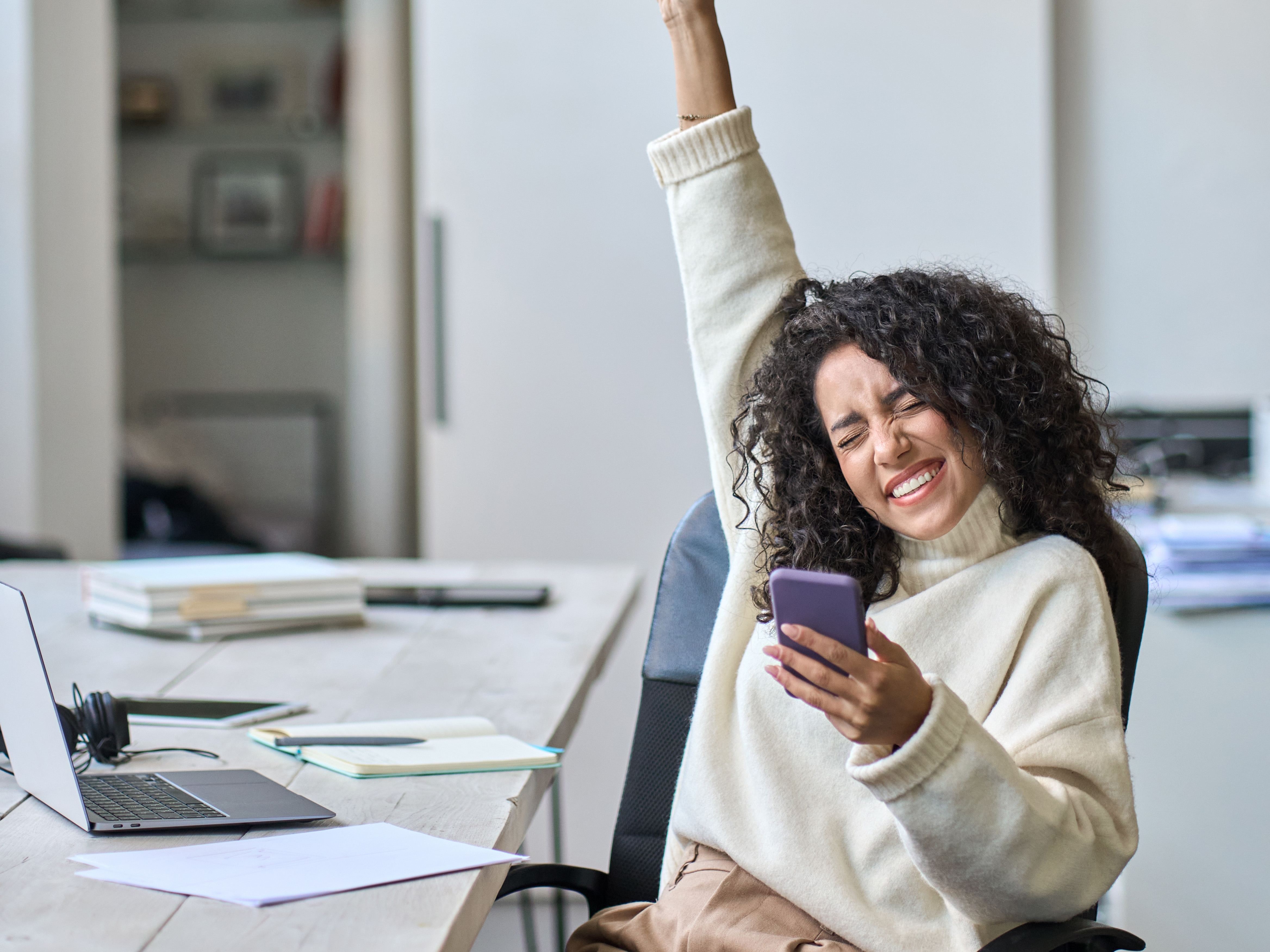 happy woman sitting at a desk holding a phone with her hand in the air