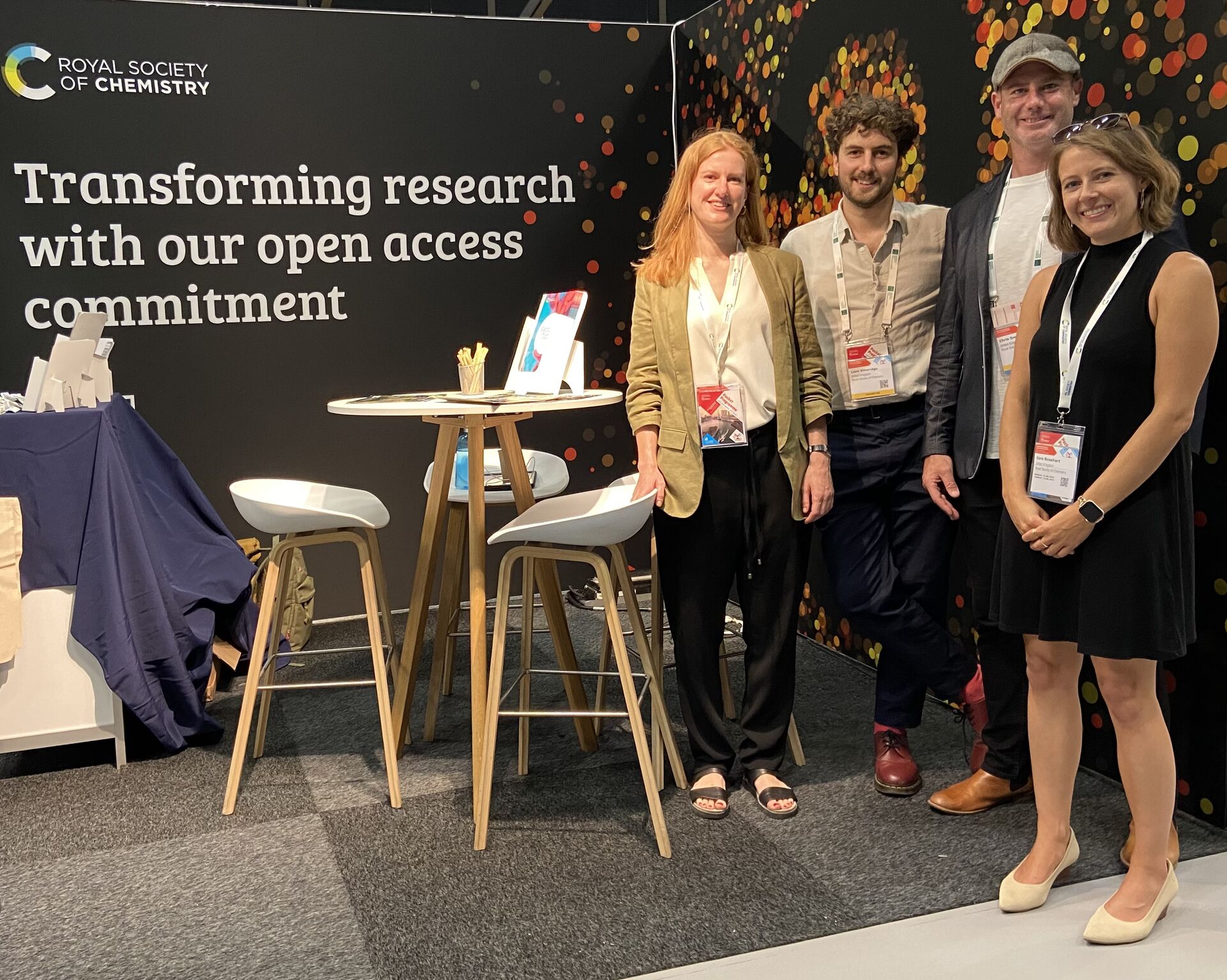 Three members of RSC staff, including Sara Bosshart (far right), stand next to a table with some stools and a banner saying "Transforming research with our open access commitment"