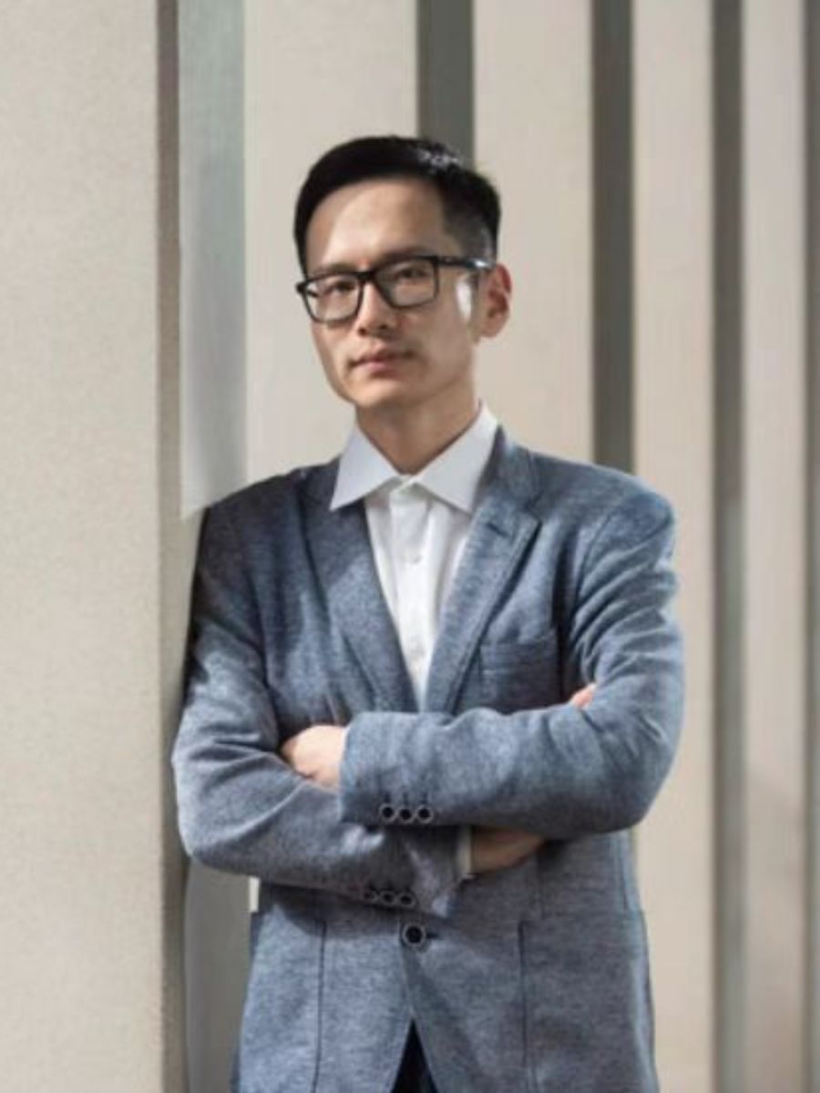 Dr Guanjie He in a suit and glasses with his arms crossed, looking at camera