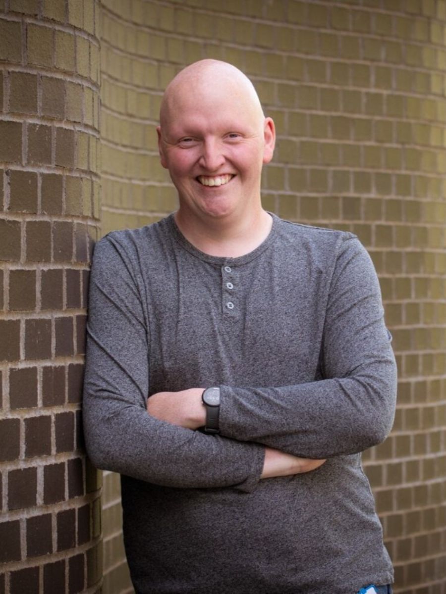 Dr Jack Woolley standing leaning on an external brick wall, smiling with arms crossed