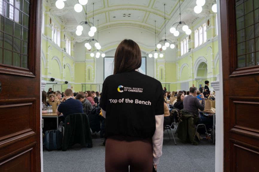 A Top of the Bench helper, facing away from the camera, stands in front of the entrance to a room full of students taking part in the event