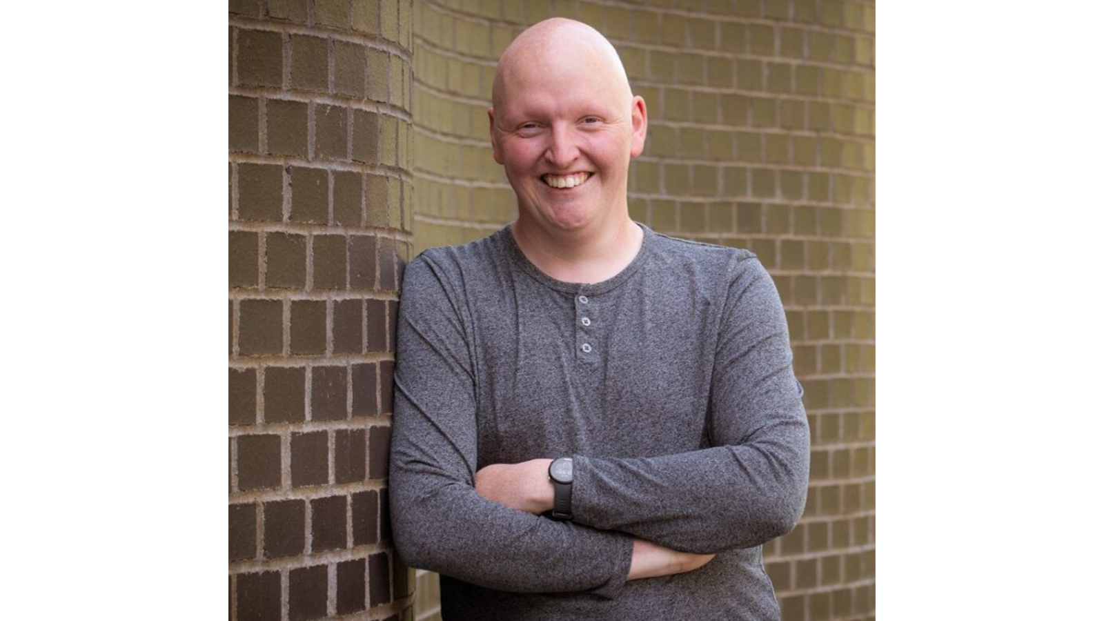 Dr Jack Woolley leaning against a brick wall outsidde, smiling with arms crossed