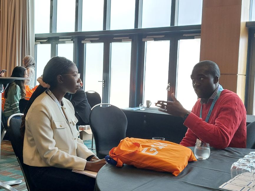 A female programme participant sits listening to a representative from GSK, while an orange GSK tote bag sits between them