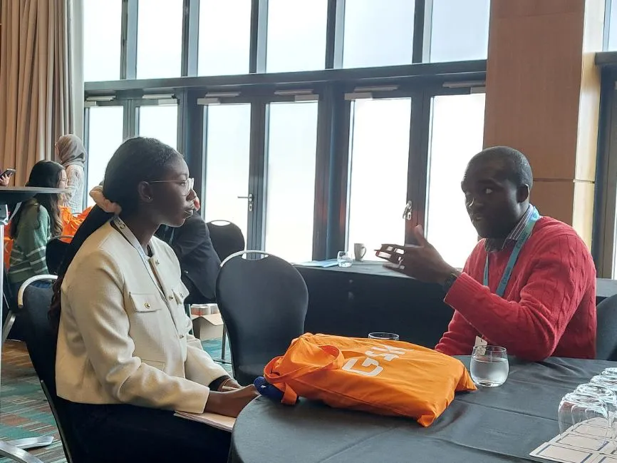 A female programme participant sits listening to a representative from GSK, while an orange GSK tote bag sits between them