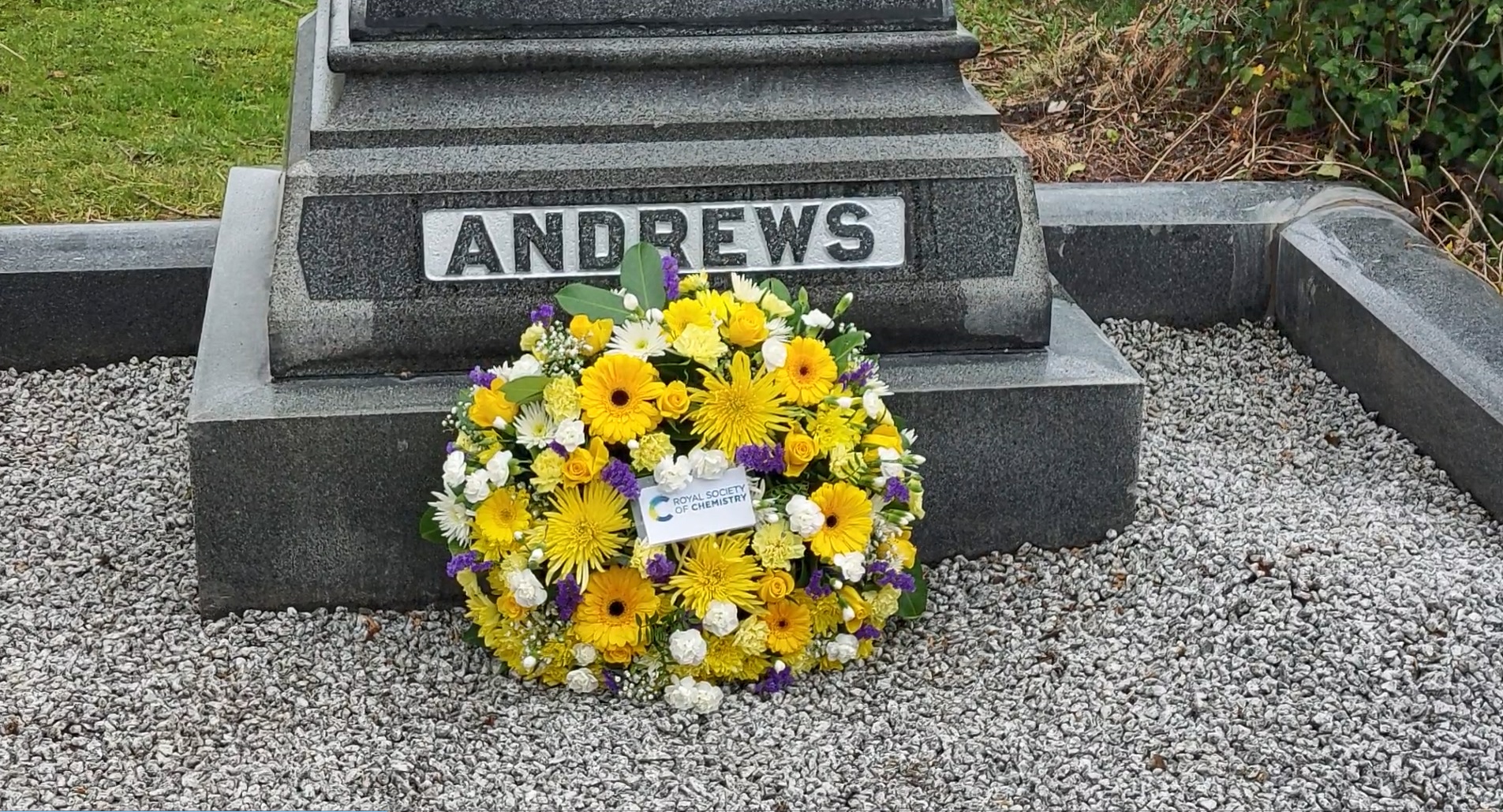 A yellow, green and blue wreath with an card with the RSC logo in the middle lies at the base of the grave
