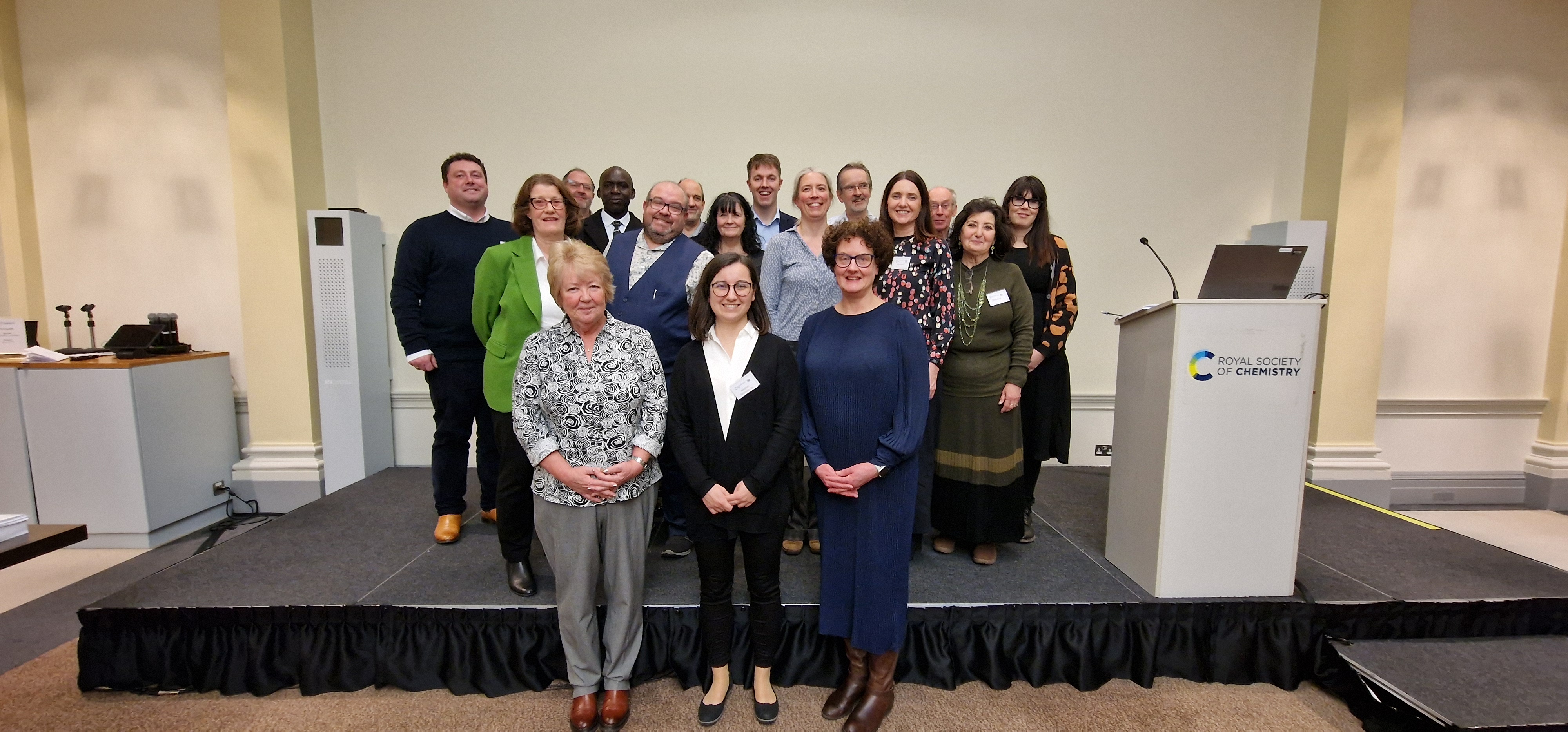 17 people pose, smiling, for a picture in front of a blank wall, with an RSC-branded podium to the right-hand side