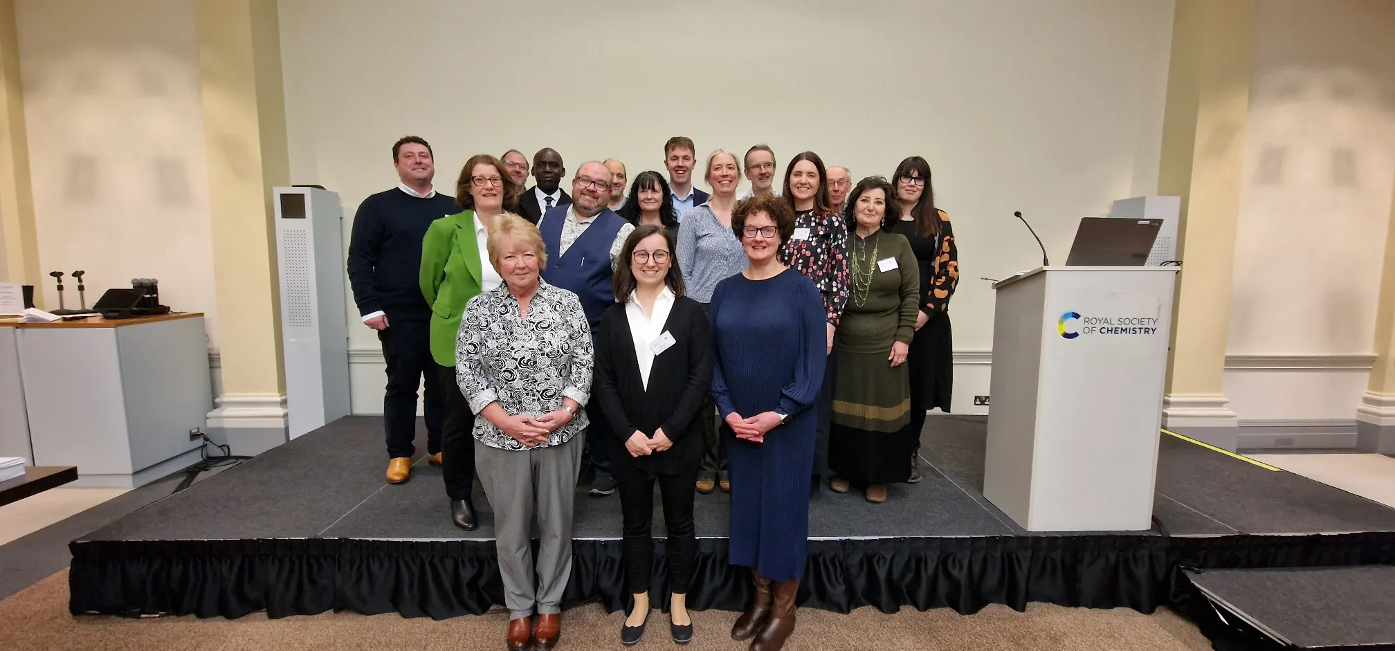 17 people pose, smiling, for a picture in front of a blank wall, with an RSC-branded podium to the right-hand side