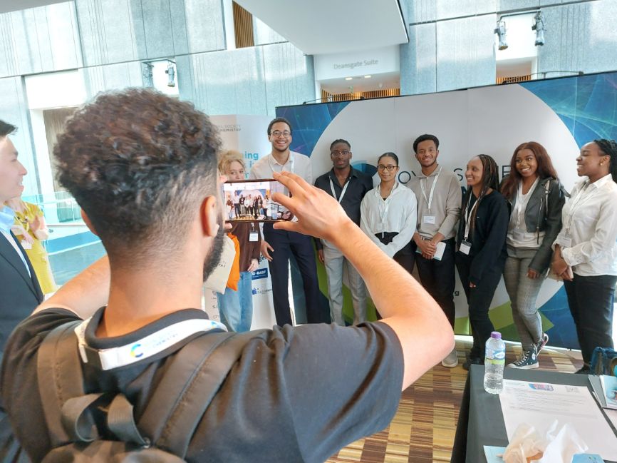 A member of the Broadening Horizons cohort takes a picture on his phone of a group of six people in front of an RSC backdrop