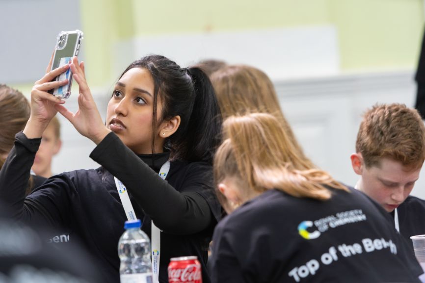 A student takes a picture on her phone while another wears a Top of the Bench-branding shirt