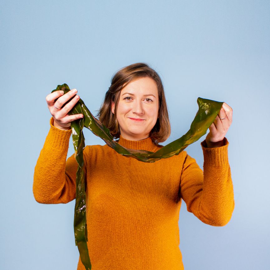 Louise Anderson, Research Director at Notpla, stands smiling and holding a long piece of seaweed Louise Anderson, Research Director at Notpla, stands smiling and holding a long piece of seaweed