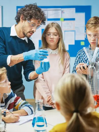 A science teacher teaches five schoolchildren in a classroom, while wearing safety goggles and doing something with glassware