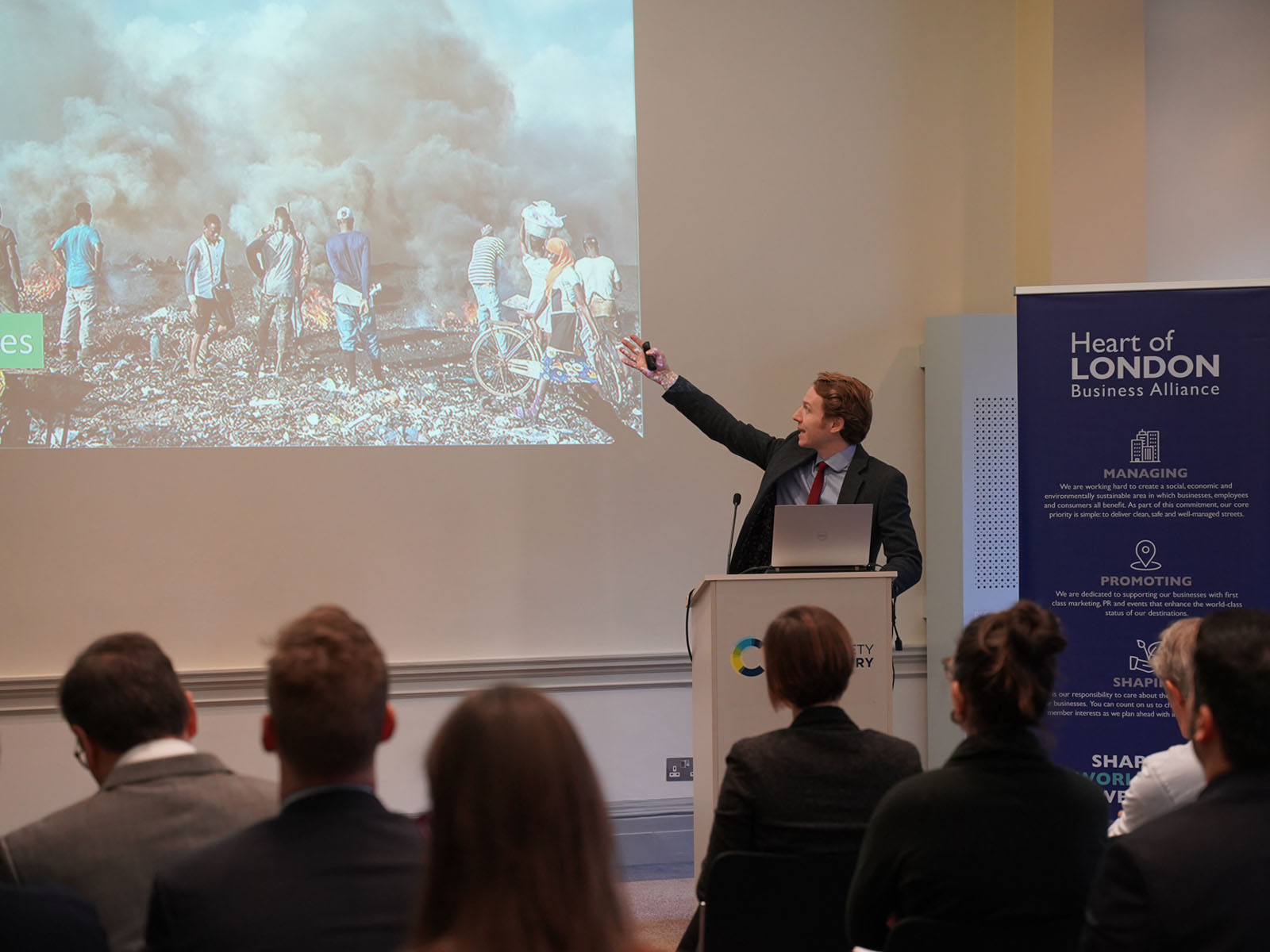 Ed Hubbard, speaking from behind a lectern, points at an image on the screen featuring a group of African men burning electronic equipment to extract the metals, with clouds of thick black smoke surrounding them