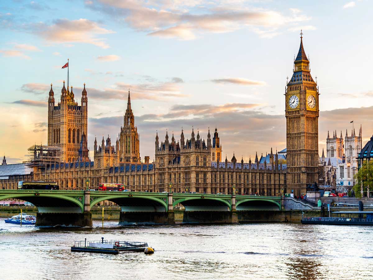 The House of Parliament stand behind the Westminster Bridge on a sunny day with a few clouds in the sky and light reflecting off the Thames.